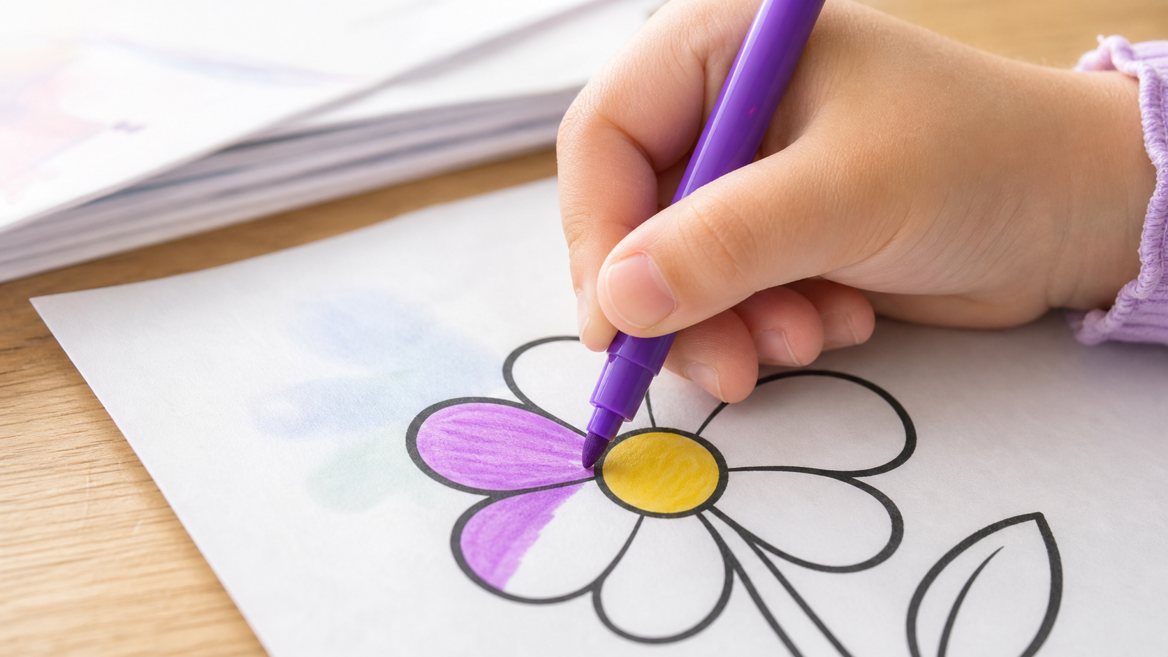 Close-up photo of a child's felt-tip marker coloring a simple bold line-art flower on a thin sheet of white paper, faint color just beginning to show through from behind the sheet, a stack of paper vi